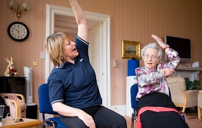 Female carer and elderly woman sitting side by side doing arm stretches