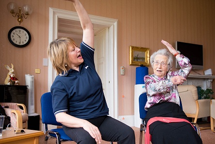 Female carer and elderly woman sitting side by side doing arm stretches