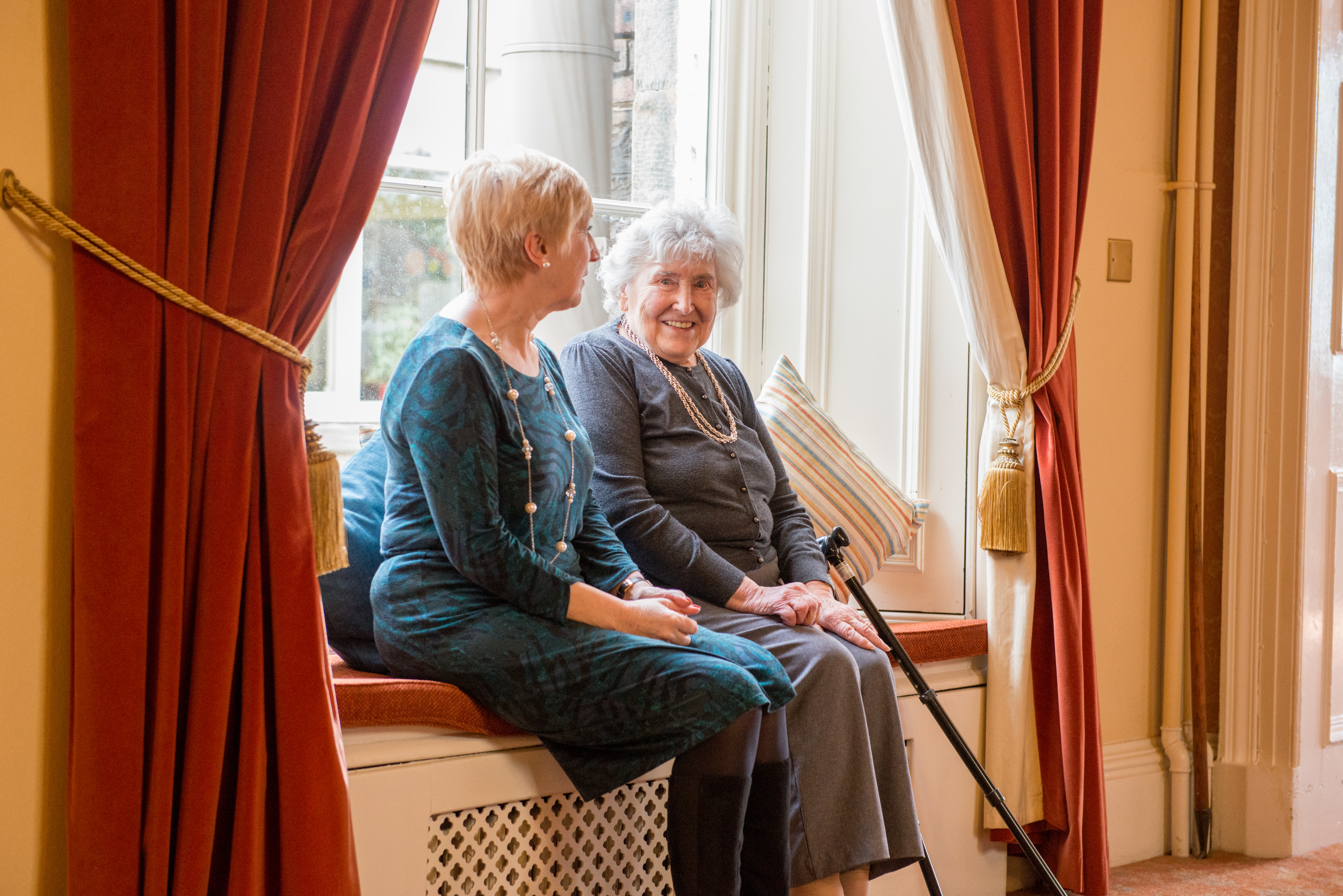 Two ladies sitting on a window seat with walking stick in view