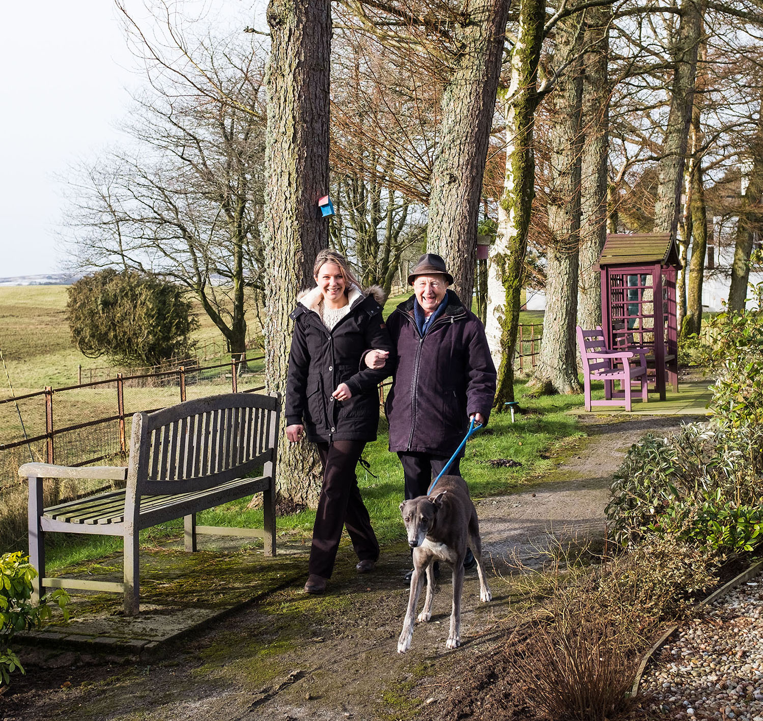 Lady and older gentleman linking arms and walking a greyhound dog in countryside 