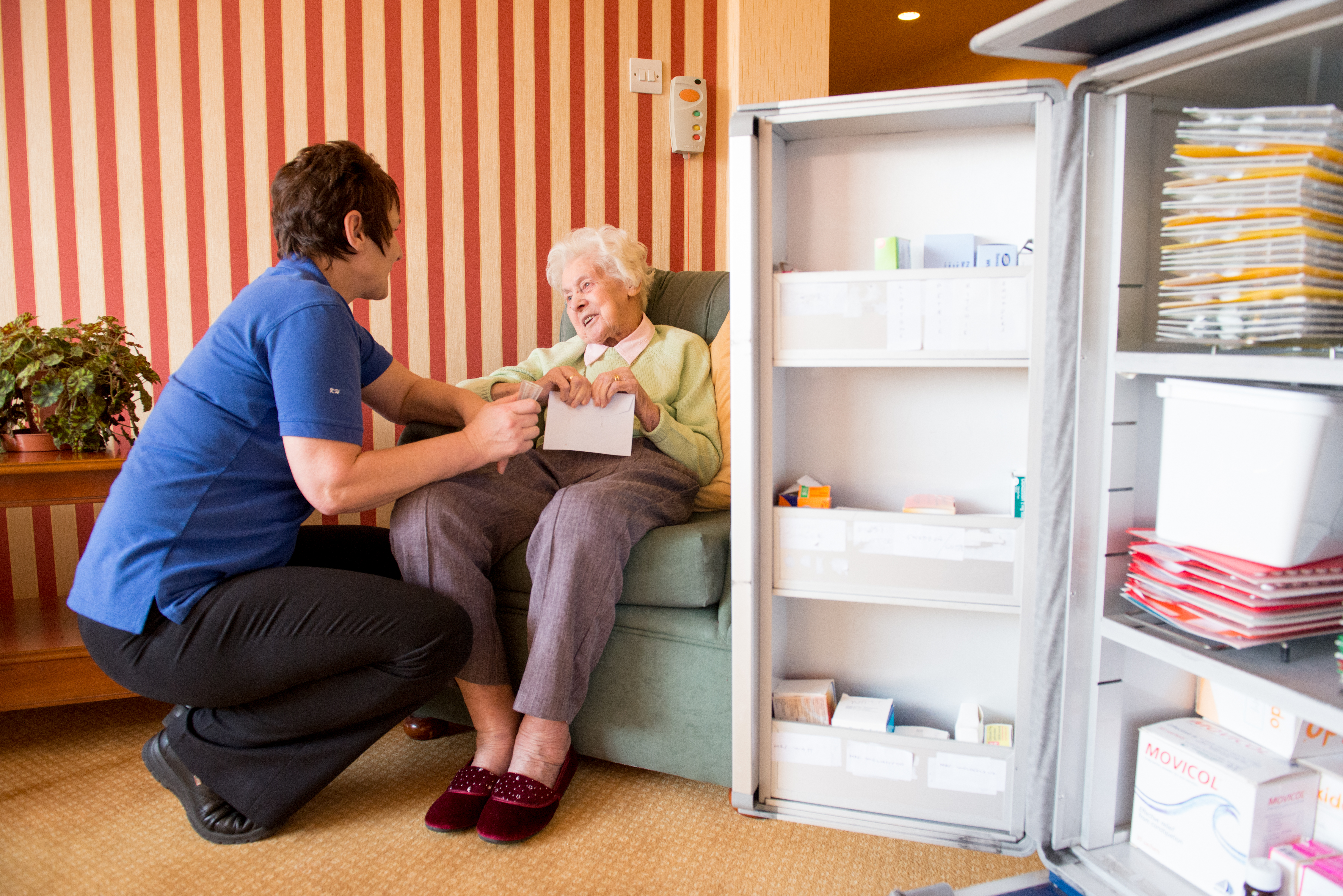 Older lady in care home speaking to a carer with a medicines fridge in the background