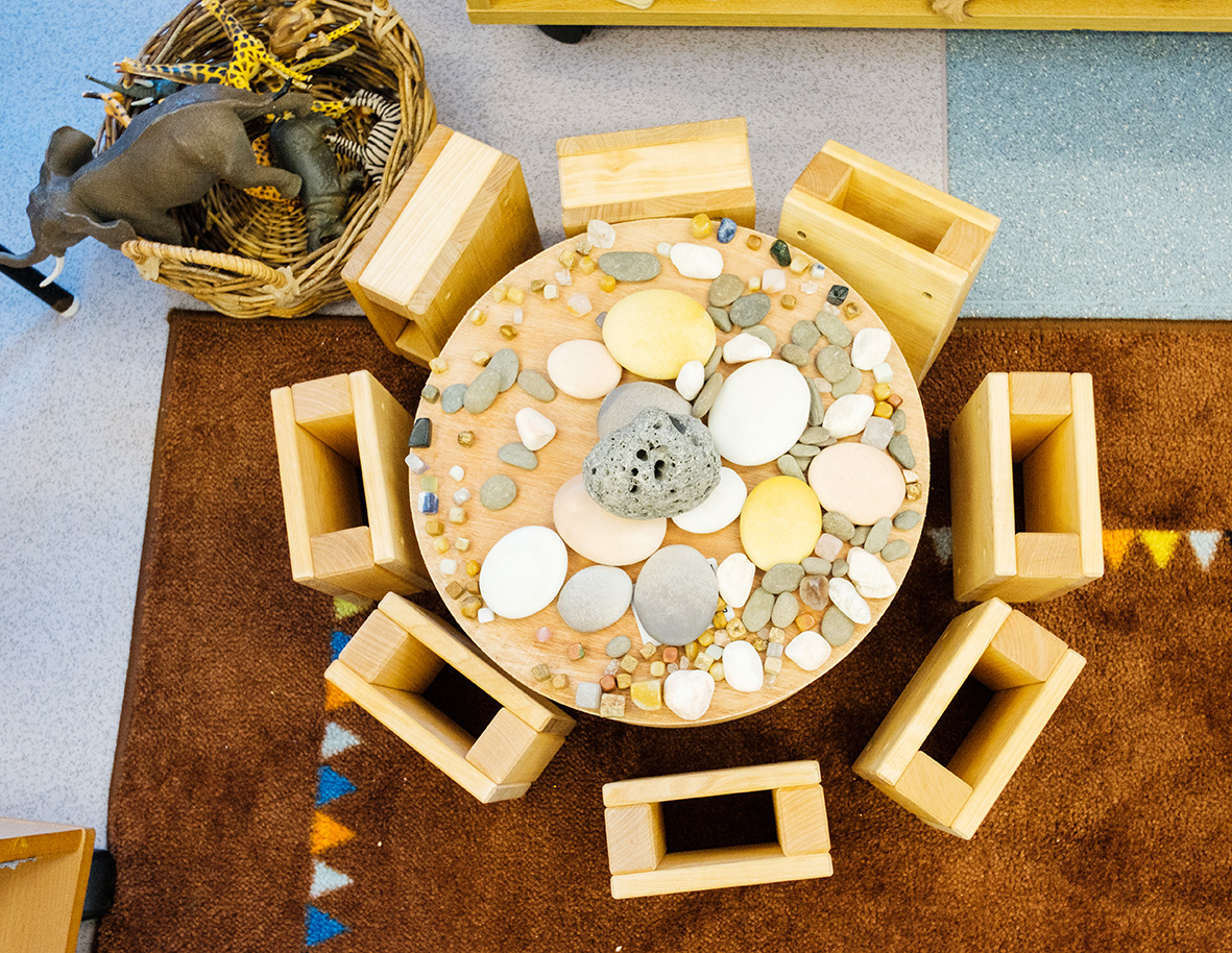 Ariel photograph of a table and wooden stools sitting on a rug. The table has different sizes of pebbles piled high and there is a basket of plastic toy animals in a basket.