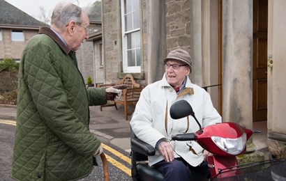 Two older men chatting outside. One is in a wheelchair and one is standing.